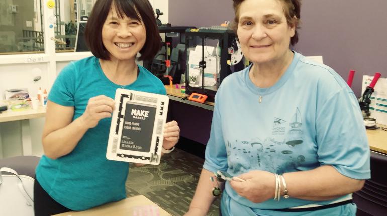 Two women posing with a picture frame that's been artfully designed with monochrome buttons and stickers.