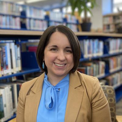 Image of a woman with short brown hair wearing a blue blouse and tan blazer, seated in front of shelves of books. 